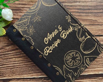 May include: Black recipe book with gold-colored text that reads "Anna's Recipe Book". The cover features gold-colored illustrations of vegetables and floral designs. The spine is black leather with brass-colored rivets.