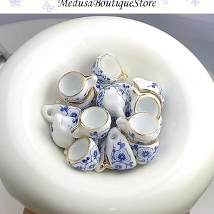 May include: A collection of miniature white porcelain teacups with blue floral patterns and gold trim, nestled in a large white bowl. The teacups have handles and are arranged in a close-up shot, showcasing their delicate design. The text "Medusa Boutique Store" is visible.