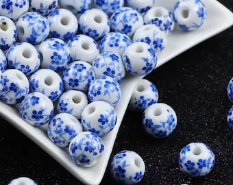 May include: White ceramic beads with blue floral patterns. The beads are round with a hole through the center, and are scattered on a white dish and a black surface. The image includes the text "Medusa Boutique Store".