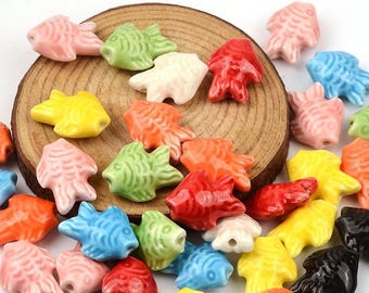 May include: A collection of colorful ceramic fish beads in various colors, including pink, green, red, yellow, orange, blue, white, and black. The beads are arranged on a wooden slice, with the text "Medusa Boutique Store" at the top.