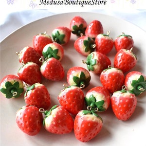 May include: A collection of 20 red and white strawberry charms with green leaves. Each charm has a gold loop for attaching to a necklace, bracelet, or other jewellery.