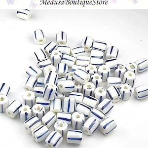 May include: A pile of white ceramic beads, each with blue stripes. The beads are cylindrical and have a glossy finish. The image includes the text "Medusa Boutique Store". These beads are ideal for jewellery making and crafts.