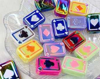 May include: A collection of iridescent playing card-shaped beads in various colors, including blue, yellow, pink, and green. Each bead features a different suit symbol: hearts, diamonds, clubs, and spades. The beads are displayed on a clear, decorative dish.