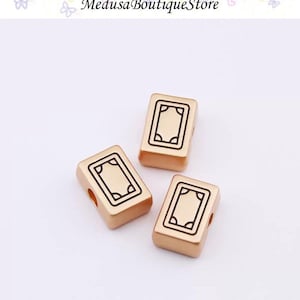 Puede incluir: Tres cuentas rectangulares doradas con un dise&ntilde;o rectangular negro. Las cuentas tienen un agujero a cada lado. El texto "Medusa Boutique Store" est&aacute; en la parte superior de la imagen.