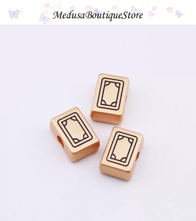 Puede incluir: Tres cuentas rectangulares doradas con un dise&ntilde;o rectangular negro. Las cuentas tienen un agujero a cada lado. El texto "Medusa Boutique Store" est&aacute; en la parte superior de la imagen.
