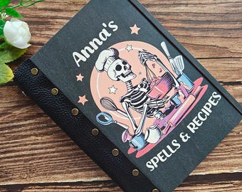 Può includere: Un ricettario nero con la scritta "Anna's SPELLS & RECIPES" e un'illustrazione di uno chef scheletro. Il libro ha un dorso in pelle nera con rivetti in ottone e una copertina in legno. L'illustrazione presenta accenti rosa, blu e bianchi.