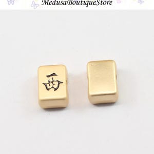 Puede incluir: Dos fichas de Mahjong doradas. Una ficha presenta un s&iacute;mbolo negro, la otra est&aacute; en blanco. Las fichas son rectangulares con bordes redondeados. El fondo es claro, y el texto "Medusa Boutique Store" es visible en la parte superior.