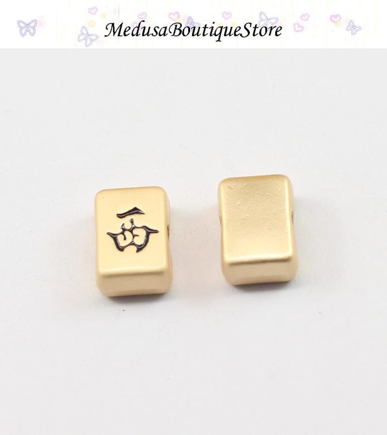 Puede incluir: Dos fichas de Mahjong doradas. Una ficha presenta un s&iacute;mbolo negro, la otra est&aacute; en blanco. Las fichas son rectangulares con bordes redondeados. El fondo es claro, y el texto "Medusa Boutique Store" es visible en la parte superior.