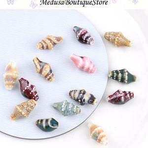May include: A collection of ceramic seashell beads in various colors, including pink, green, brown, and white. The beads have a glossy finish and are arranged on a white surface. The text "Medusa Boutique Store" is visible at the top.