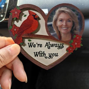 Pode incluir: Enfeite de madeira em forma de coração com uma foto de uma mulher e um pássaro cardeal vermelho. O enfeite inclui o texto "We're Always With You" e detalhes florais vermelhos. O enfeite é segurado por uma mão.