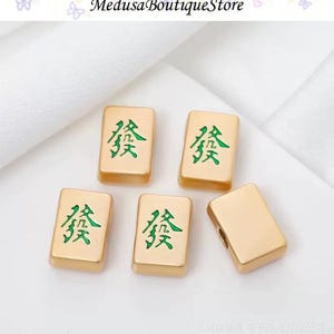 Puede incluir: Cinco fichas de Mahjong doradas con caracteres chinos verdes. Las fichas son rectangulares y tienen una superficie lisa y reflectante. El texto "Medusa BoutiqueStore" est&aacute; en la parte superior.