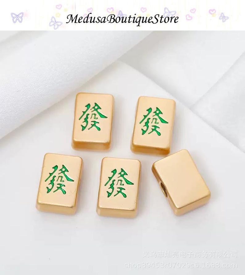 Puede incluir: Cinco fichas de Mahjong doradas con caracteres chinos verdes. Las fichas son rectangulares y tienen una superficie lisa y reflectante. El texto "Medusa BoutiqueStore" est&aacute; en la parte superior.
