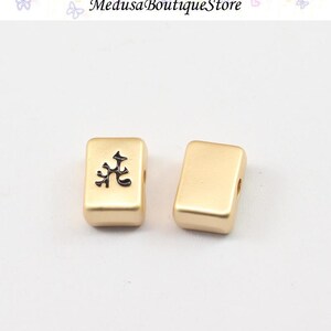 Puede incluir: Dos cuentas rectangulares doradas. Una cuenta tiene un s&iacute;mbolo negro. Las cuentas tienen agujeros para ensartar. El texto "Medusa Boutique Store" est&aacute; en la parte superior de la imagen.