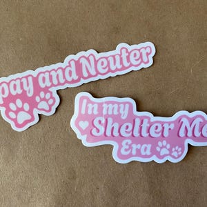 May include: Two pink stickers with white text. The first sticker says "Spay and Neuter" with two paw prints. The second sticker says "In my Shelter Era" with a heart and two paw prints.