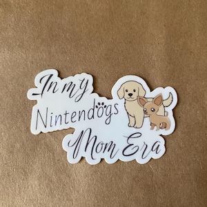 May include: White sticker with the text "In my Nintendogs Mom Era" and a cartoon image of two dogs. The larger dog is a golden retriever and the smaller dog is a chihuahua.