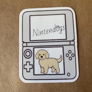 May include: A gray and white sticker of a Nintendo DS with the text "Nintendogs" on the top screen. The bottom screen shows a cartoon image of a golden retriever puppy.