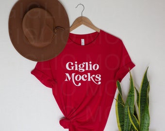 May include: Red t-shirt with the text "Giglio Mocks" in white on a white hanger. A brown hat and a green plant are in the background.
