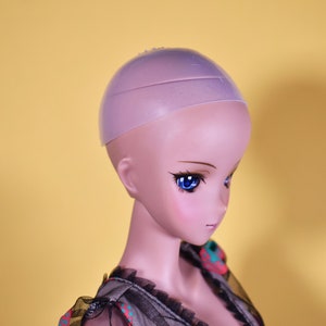 May include: A doll head with blue eyes and a pink complexion, wearing a light purple wig cap. The doll is wearing a black and patterned top with a ruffled neckline. The background is a solid yellow.