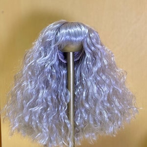 May include: A long, curly, silver-grey wig for a doll. The wig is on a stand and has a center part.