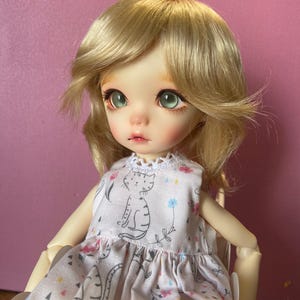 May include: A doll with blonde hair wearing a pink dress with a cat print. The doll has green eyes and is sitting on a white chair.