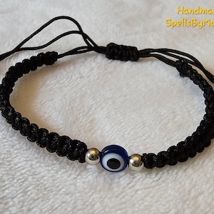 May include: Black braided cord bracelet featuring a blue and white evil eye bead and silver-colored beads. The bracelet is adjustable with sliding knots and has the text "Handmade by SpellsByMoonlight".