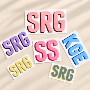 May include: A collection of six rectangular stickers featuring bold, block-letter monograms. The stickers display the letters SRG in pink, purple, yellow, and green, and the letters SS in pink, and KGE in blue, all with a distinct drop shadow effect.