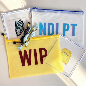 May include: Two mesh zipper pouches, one yellow with the text WIP and one white with the text NDLPT, containing two needlepoint canvases featuring an oyster and a yellow sweater with the text CAPE COD, plus several skeins of embroidery floss.