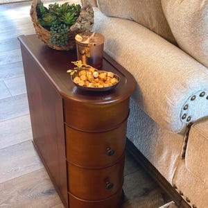 May include: A narrow, dark wood end table with two rounded drawers featuring small dark knobs. On top sits a woven basket filled with green artificial succulents, a cylindrical candle with a metallic lid, and a small bowl of gold-wrapped candies.