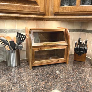 May include: A wooden bread box with a clear glass front and a hinged top. The bread box is made of light brown wood and has a brass handle. It is sitting on a brown countertop.