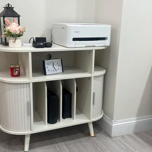 May include: A white, multi-tiered storage cabinet with a printer, clock, and decorative items. The cabinet has a curved design with vertical slatted doors and a printer on top. A black lantern and a small floral arrangement are also on the top shelf.