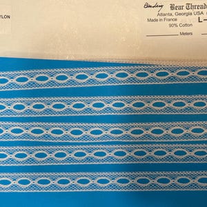 May include: A roll of white lace trim with an openwork design, set against a vibrant blue fabric. The trim is made of 90% cotton and 10% nylon. The label indicates it was made in France.