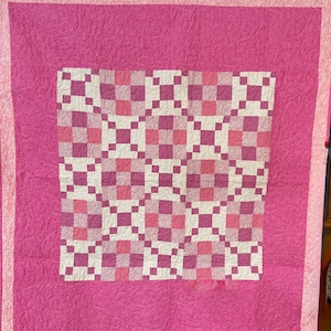 May include: A handmade quilt in shades of pink and white. The center features a checkerboard pattern of squares in varying shades of pink and white. The quilt is framed by a solid pink border and a patterned pink edge.