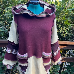 May include: A burgundy and cream knitted sweater with a ruffled hood and patchwork details. The sweater has a relaxed fit and a unique, layered design.
