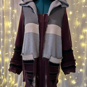 May include: A long, burgundy zip-up jacket with a gray hooded top, featuring a patchwork design with white and gray stripes and black accents.