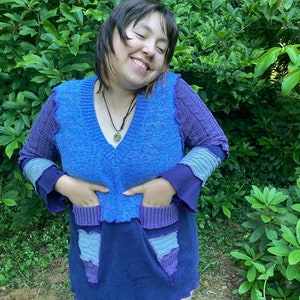May include: A woman wearing a blue and purple patchwork sweater dress with a v-neck and long sleeves. The dress has pockets and a scalloped hem.