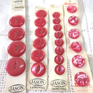 May include: Three cards of pink plastic buttons with the brand name "Jason Laundwell" printed on each card. The buttons are in various sizes and shapes, including round, oval, and a swirl design.