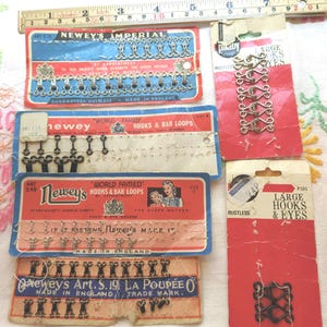 May include: Assortment of vintage sewing notions, including hooks and eyes, bar loops, and a hook and eye tape. The packaging is red, white, and black with the brand name "Newey's" and the text "World Famed" and "Made in England".