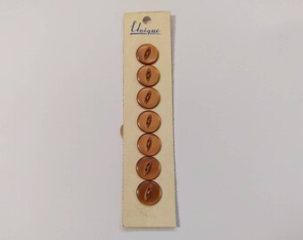 Puede incluir: Una tarjeta con siete botones marrones de forma ovalada con un diseño central alargado. Los botones están sujetos a una tarjeta vintage con la palabra "Unique" impresa en la parte superior. Los botones tienen un acabado brillante.
