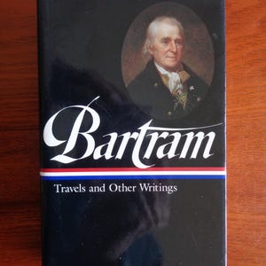 May include: A hardcover book with a black cover featuring the name "Bartram" in white script. The book includes a portrait of a man and the text "Travels and Other Writings" in white.