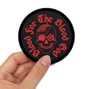 Puede incluir: Una pegatina redonda negra con texto rojo que dice "Blood for the Blood God" y una calavera roja con un solo ojo.
