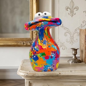 Peut inclure: Un vase fantaisiste et coloré avec un design unique en forme de canard. Le vase présente un effet de peinture éclaboussée vibrante dans les tons de bleu, orange, jaune et rose. Il a des yeux blancs et globuleux et repose sur une table blanche ornée.