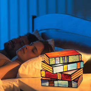 Könnte beinhalten: Eine Tischlampe aus Buntglas in Form eines Stapels aus vier Büchern. Die Lampe besteht aus leuchtend roten, grünen, gelben und blauen Glaselementen mit dunklen Metallrahmen. Sie strahlt ein warmes Licht aus dem Inneren der Glasbücher aus.