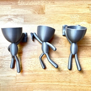 May include: Three gray, stylized planter figures with a unique design. Each figure has a rounded head and a body with arms and legs, appearing to hold a small rectangular piece. The planters are designed to be mounted on a wall.