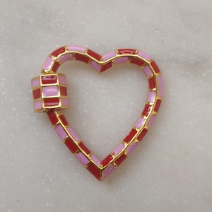 May include: A heart-shaped charm with a pink and red enamel design. The charm is made of gold-toned metal and has a square-shaped clasp.