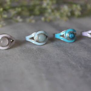 May include: Four silver rings with different colored enamel and gemstone accents. The rings feature a stylized eye design with a white enamel border. The gemstones are a clear quartz, a light blue aquamarine, a turquoise, and a purple amethyst.