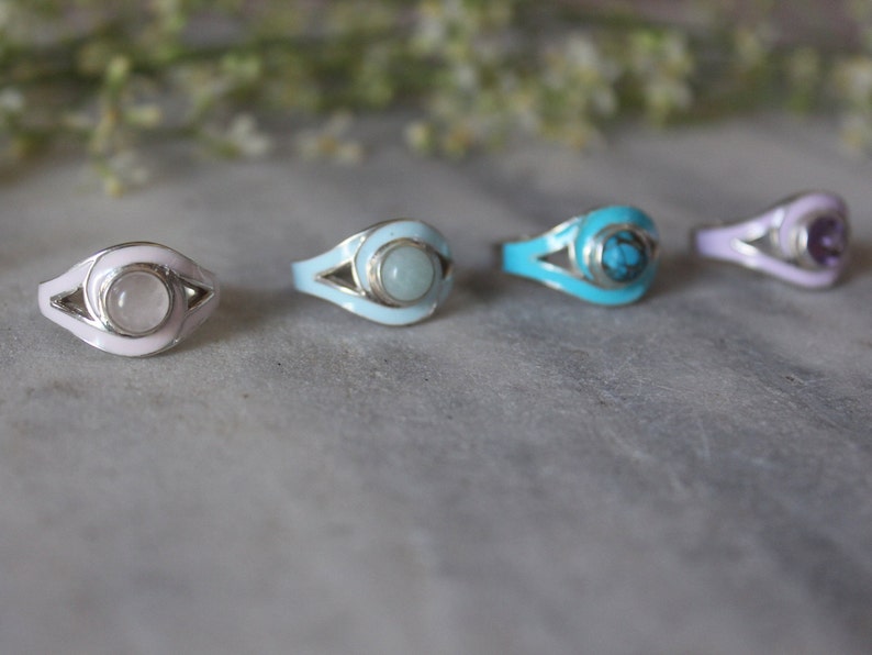 May include: Four silver rings with different colored enamel and gemstone accents. The rings feature a stylized eye design with a white enamel border. The gemstones are a clear quartz, a light blue aquamarine, a turquoise, and a purple amethyst.