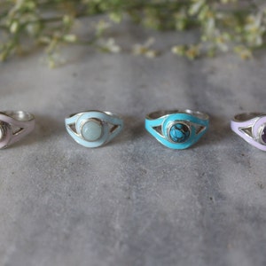 May include: Four silver rings with different colored enamel and gemstones. The rings feature a curved design with a gemstone in the center. The gemstones are pink, light blue, turquoise, and purple.