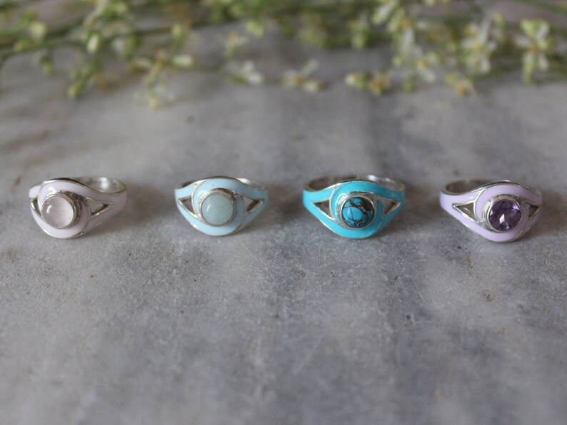 May include: Four silver rings with different colored enamel and gemstones. The rings feature a curved design with a gemstone in the center. The gemstones are pink, light blue, turquoise, and purple.