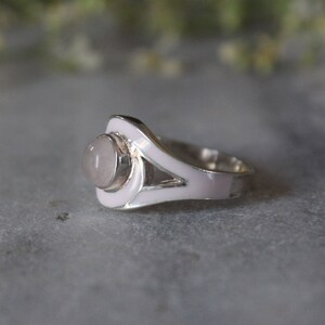 May include: A silver ring with a pink enamel design and a pale pink gemstone in the center.