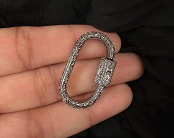 May include: A silver-colored, oval-shaped carabiner with intricate floral and scrollwork engravings. The locking mechanism is rectangular and also features detailed patterns. The carabiner is held in a hand against a dark background.